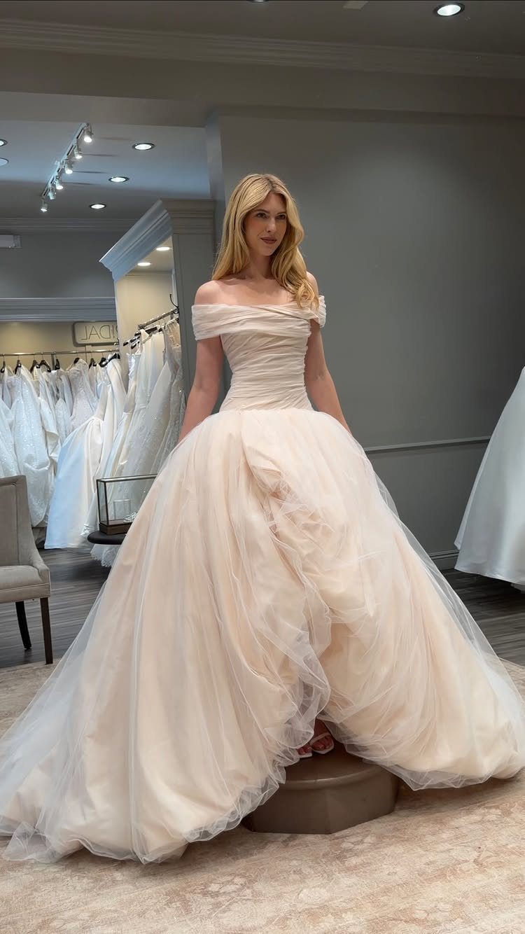 Pink Ballgown Wedding Dress: 8 Ideas with Bows and Feathers 6 Woman wearing a pale pink off-the-shoulder ballgown wedding dress in a bridal boutique.