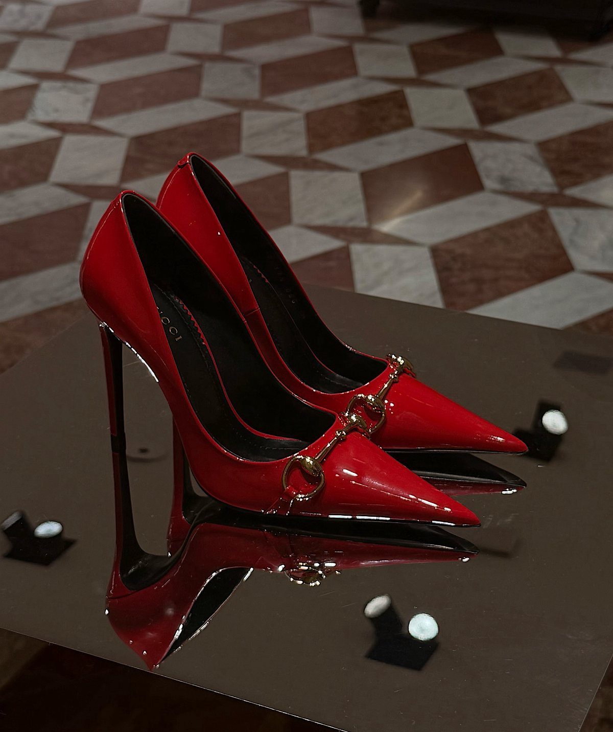 Red pointed-toe high heel pumps with gold chain details on a reflective surface.
