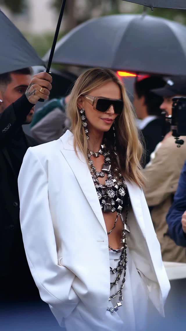 Woman in white oversized blazer and chunky silver jewelry posing outdoors with umbrellas.