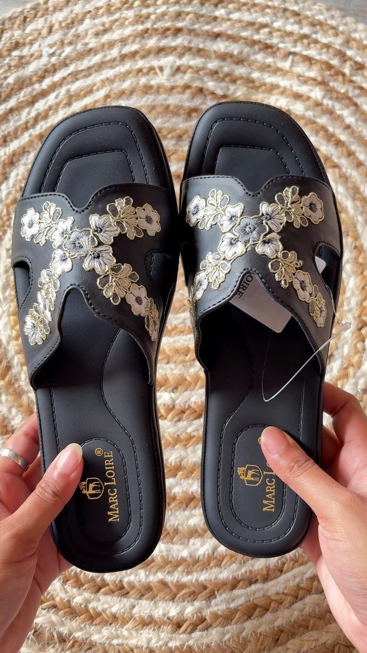 Black open-toe mules with gold floral embroidery held over a woven straw mat.