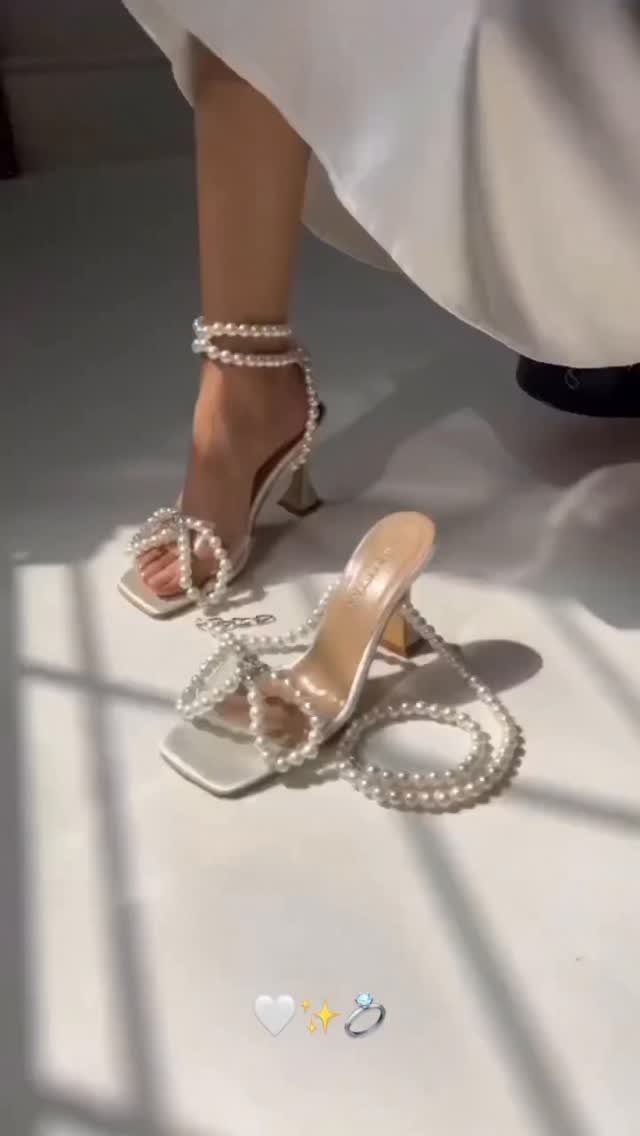 White pearl-embellished high-heeled sandals worn with a long white dress on a floor.