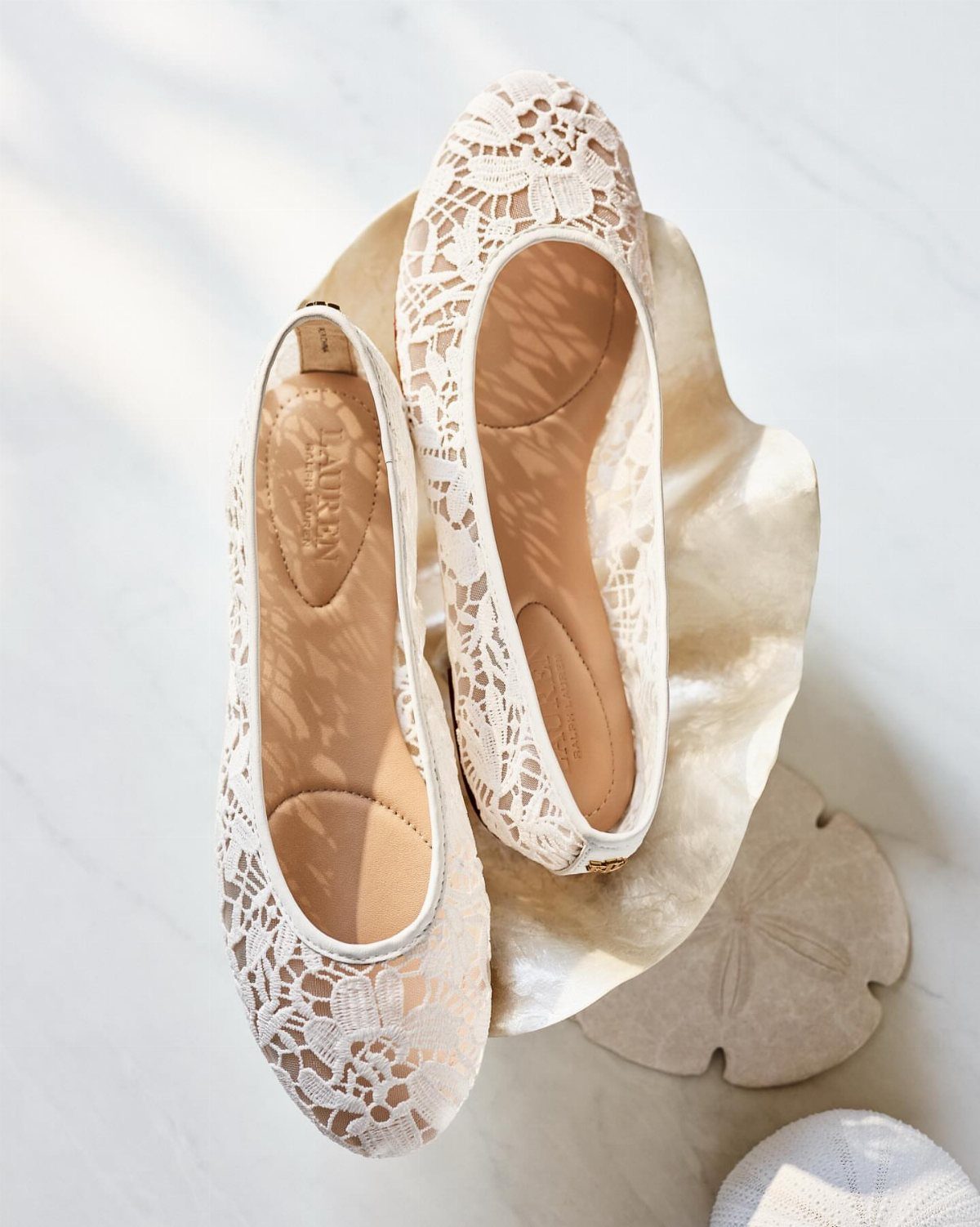 Ivory lace pointed-toe flat shoes on a white marble surface with a shell.