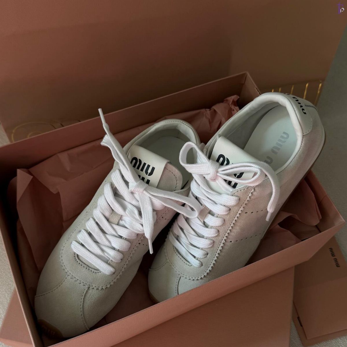 White suede Miu Miu sneakers inside a pink shoebox with tissue paper.