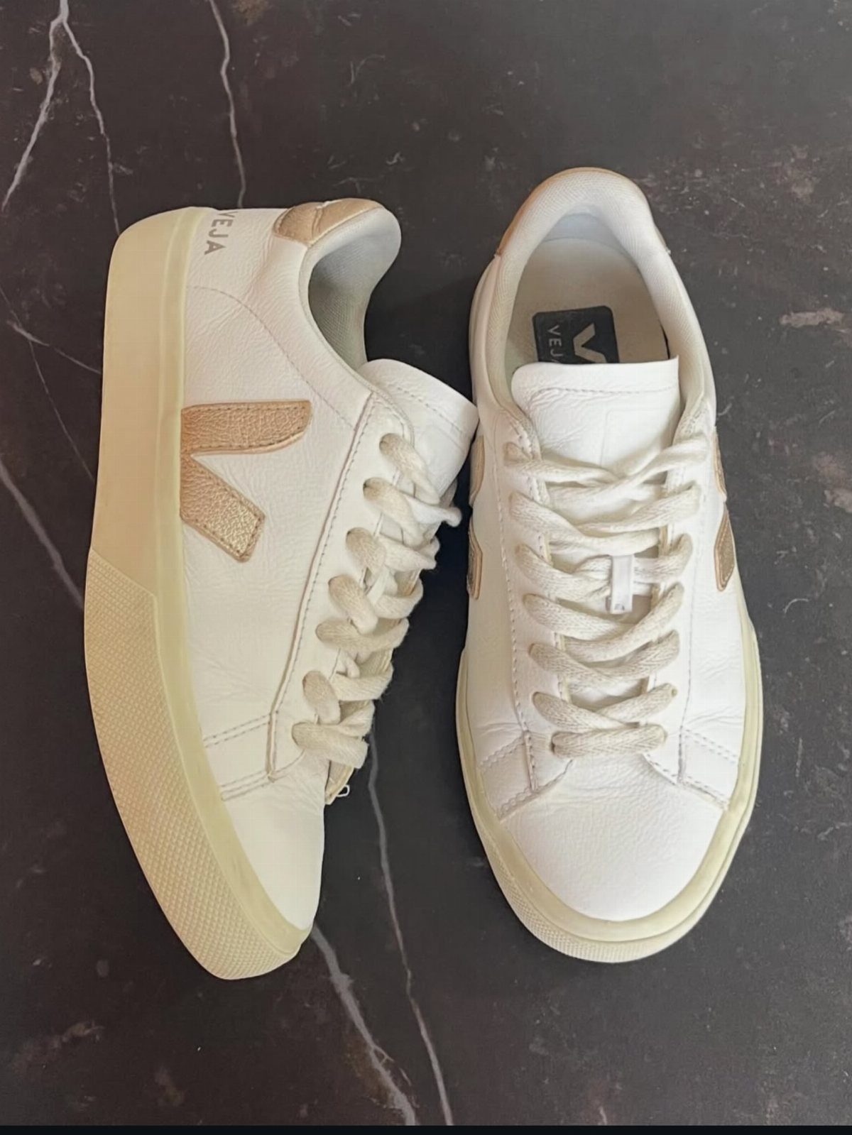 White Veja V-90 sneakers with gold logos on a dark marble background.
