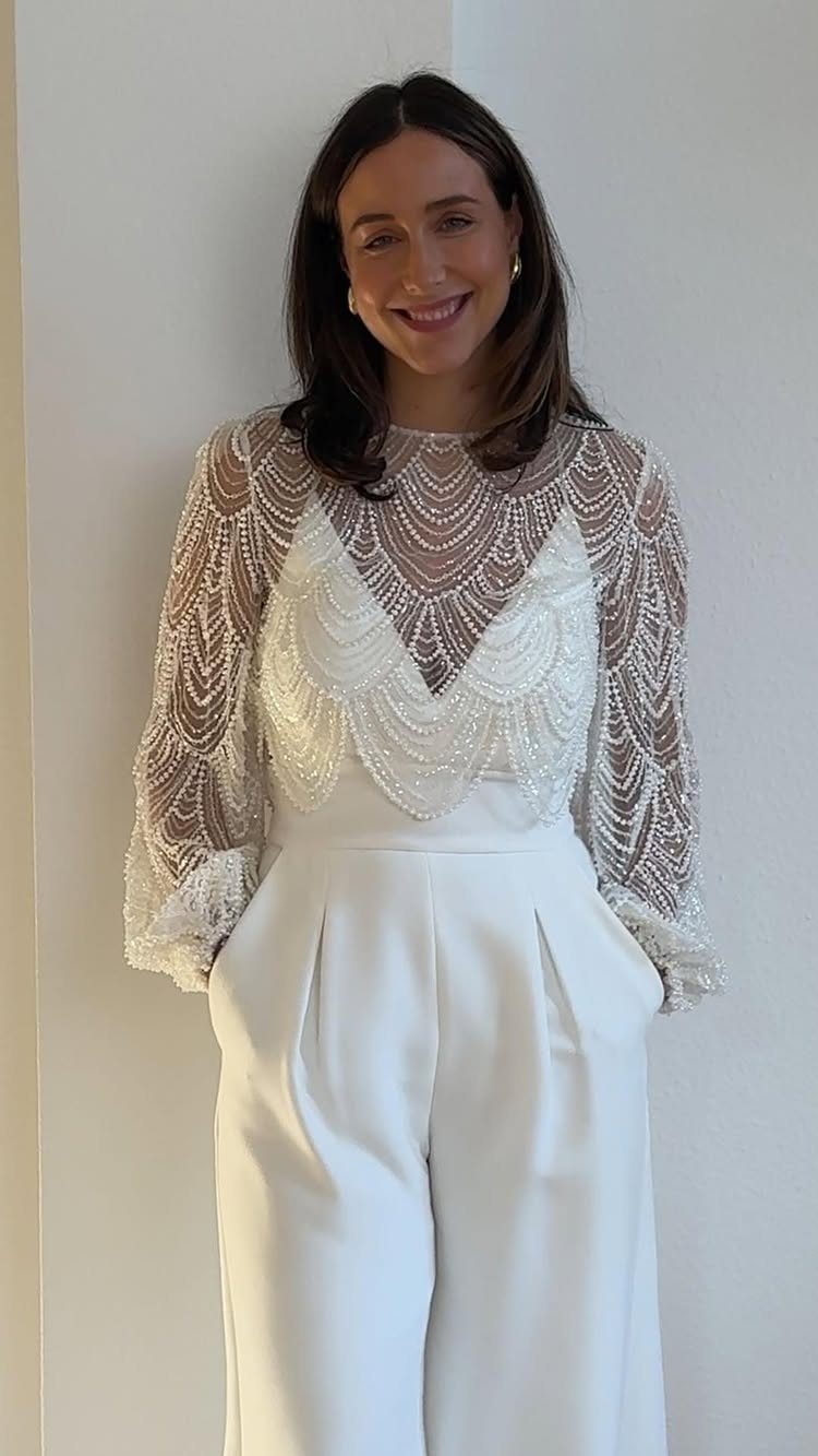 10 Wedding After Party Outfit Ideas for Guests 3 Woman wearing a white beaded long-sleeve jumpsuit with wide-leg trousers posing indoors.