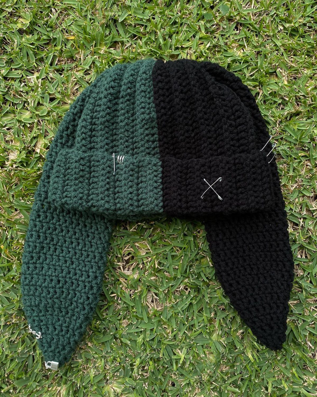 Two-toned green and black crocheted beanie with earflaps resting on green grass.