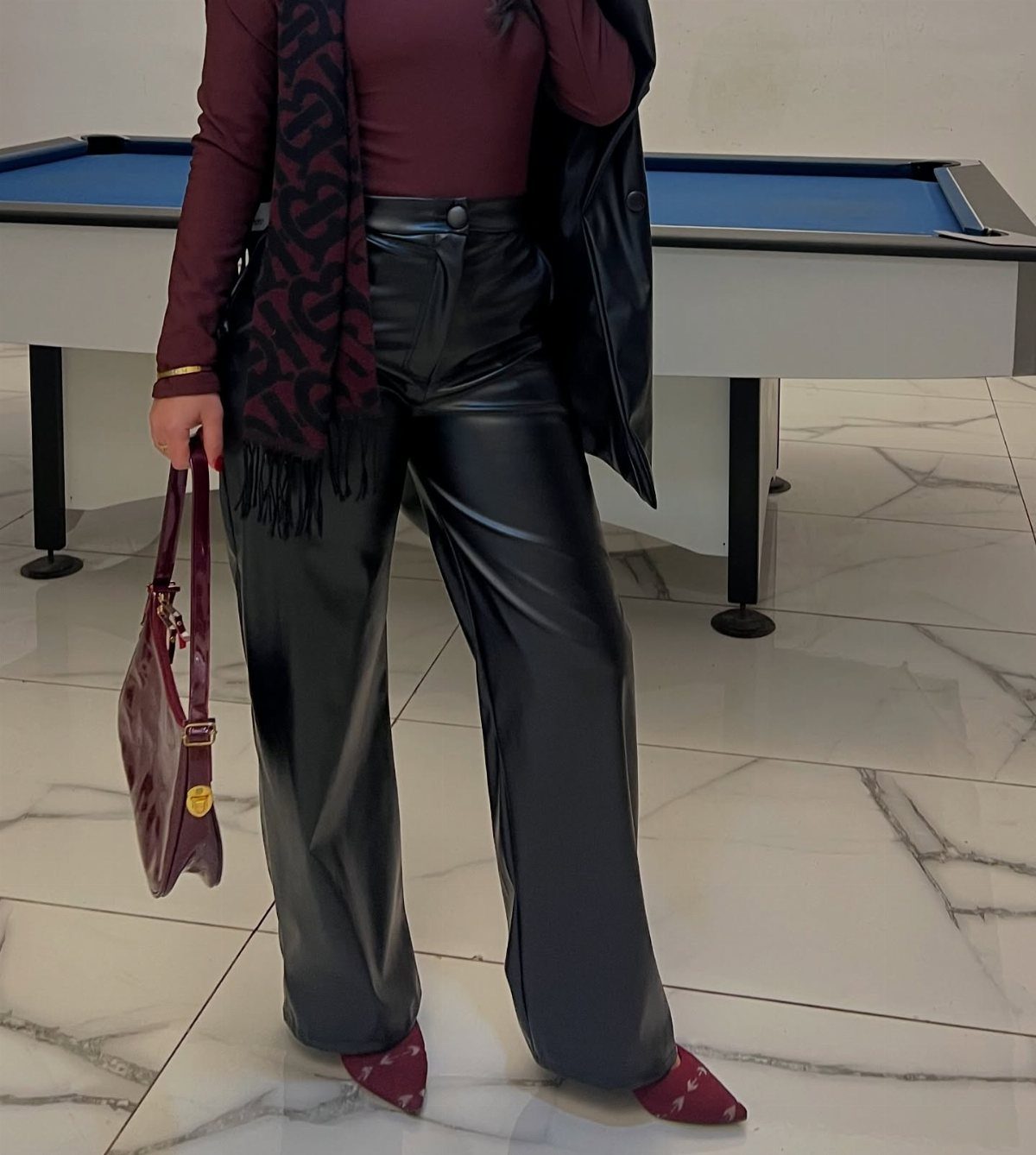 Person in burgundy top, black leather trousers, and scarf standing by pool table