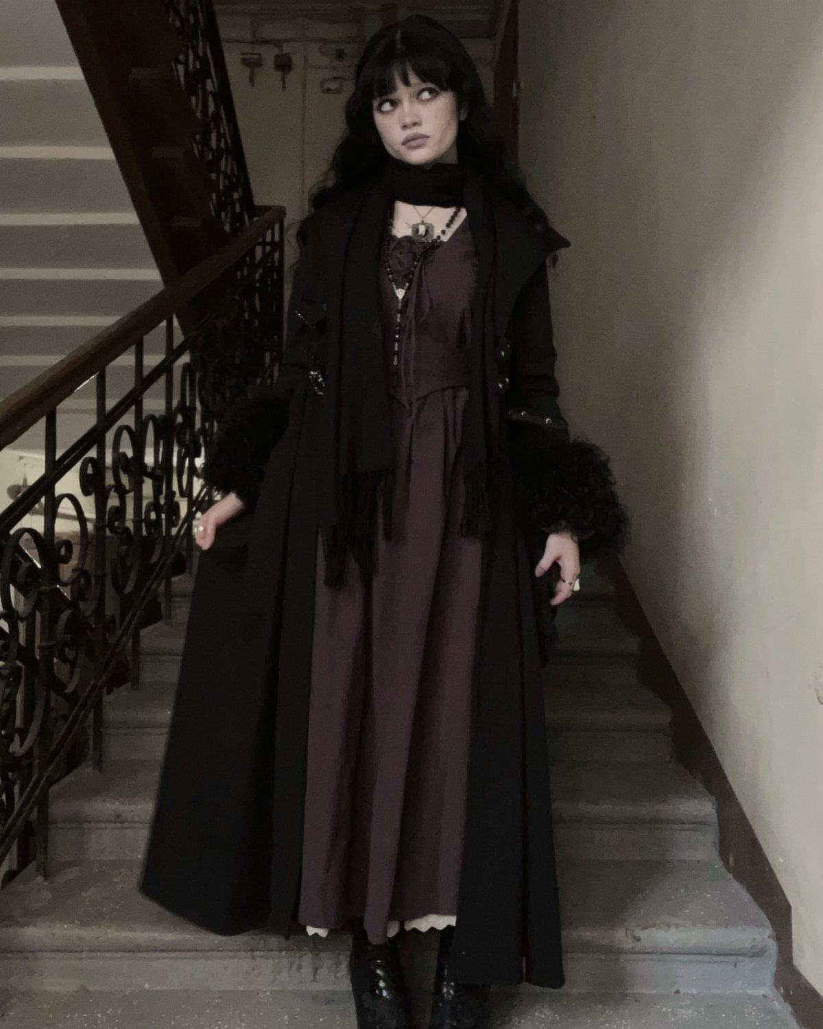 Woman in a long black coat and purple dress standing on stairs.